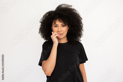 Worried young woman biting fingernail. Caucasian female model with afro hairstyle and brown eyes in black T-shirt looking nervous hearing something unpleasant. Anxiety, trouble concept