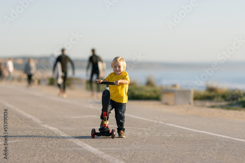 Smiling little boy stands on the road with a scooter