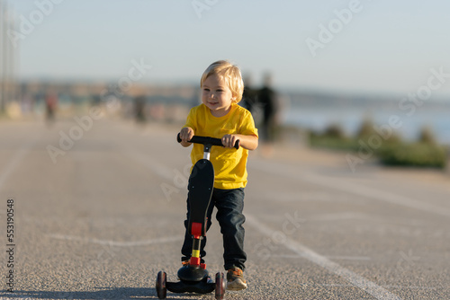 Smiling little boy stands on the road holding by scooter