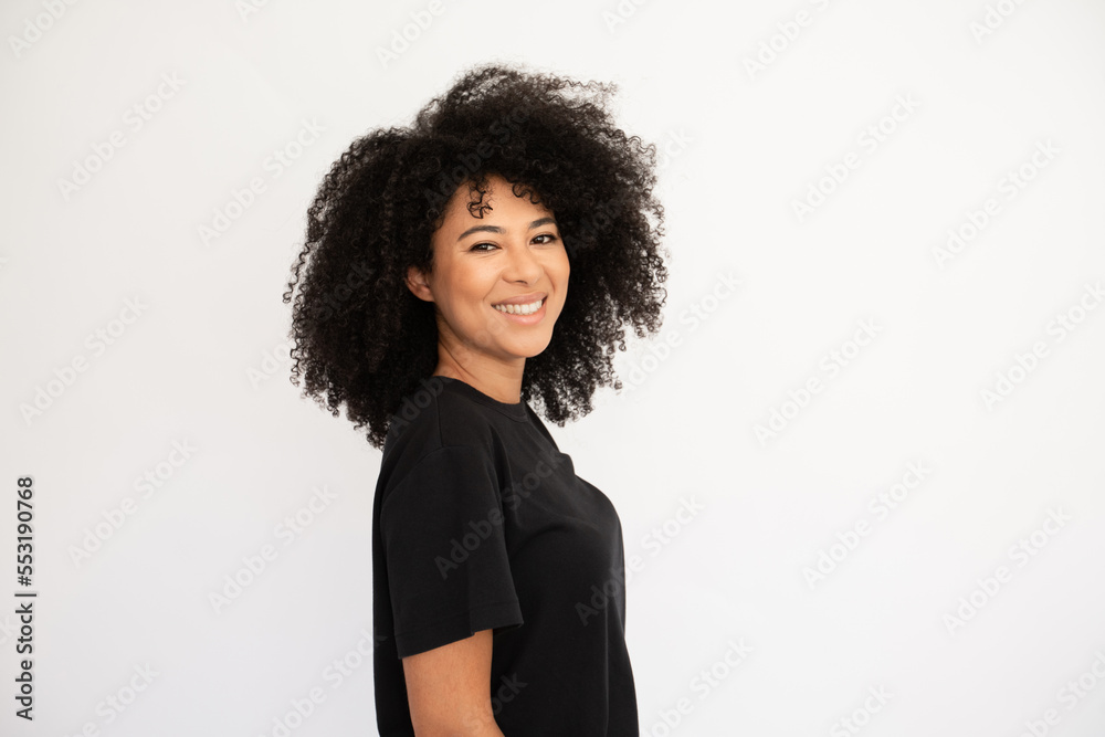 Joyful young woman smiling. Hispanic female model with afro hairstyle and brown eyes in casual clothes smiling from joy standing sideways. Emotion, happiness concept Joyful young woman smiling. Hispanic female model with afro hairstyle and brown eyes in casual clothes smiling from joy standing sideways. Emotion, happiness concept
