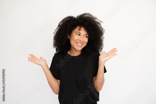 Joyful young woman shrugging throwing up hands looking away. Caucasian female model with afro hairstyle and brown eyes in casual clothes looking confused spreading hands. Uncertainty, doubt concept © KAMPUS