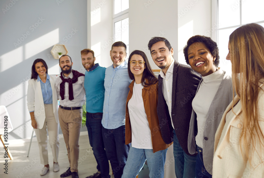 Support and union. Portrait of happy multiracial business colleagues and friends hugging and smiling at camera. Side view of happy diverse corporate staff standing in row in bright modern office.