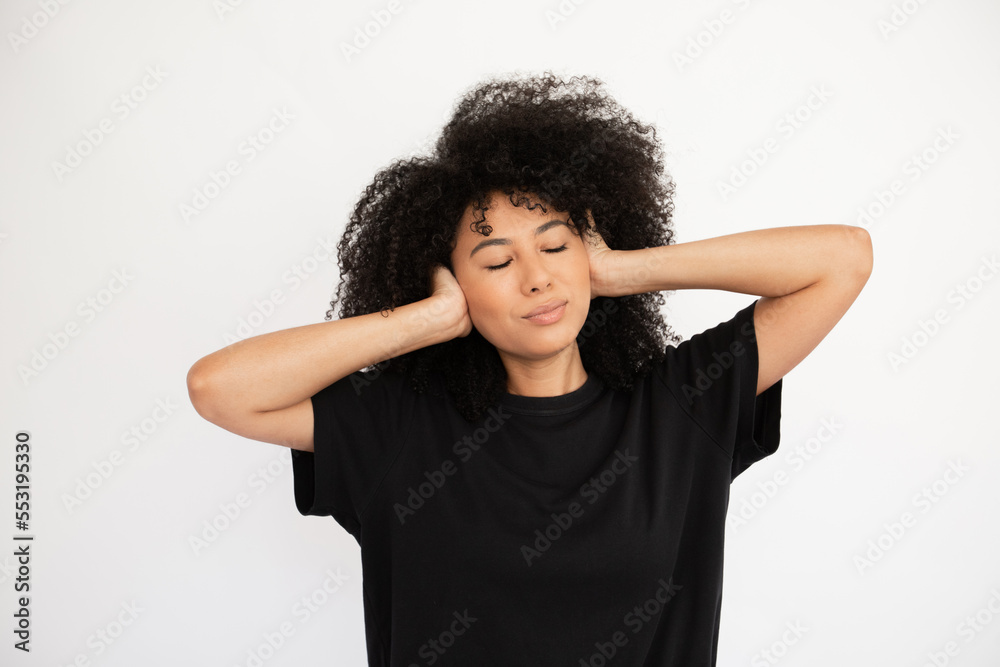 Dreaming young woman with closed eyes covering her ears with hands. Hispanic female model with afro hairstyle and closed eyes in black T-shirt closing her ears concentrating. Silence, privacy concept Dreaming young woman with closed eyes covering her ears with hands. Hispanic female model with afro hairstyle and closed eyes in black T-shirt closing her ears concentrating. Silence, privacy concept