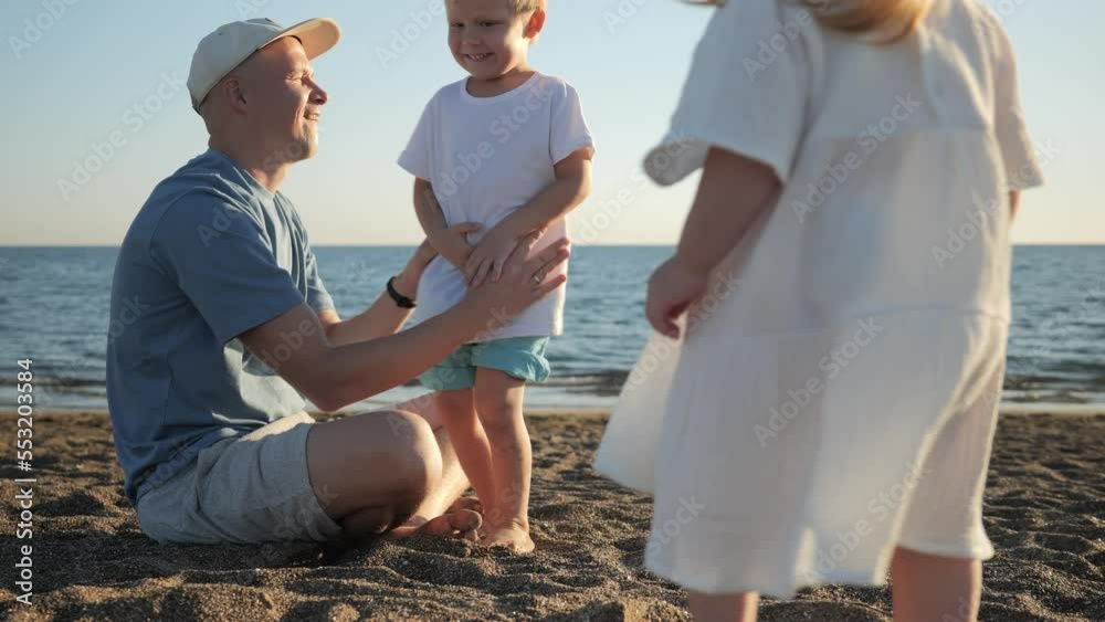 A little boy and girls resort to their dad and sit on his lap on the beach near the sea during sunset. Happy childhood by the sea. Brother and sister with their dad. High quality 4k footage