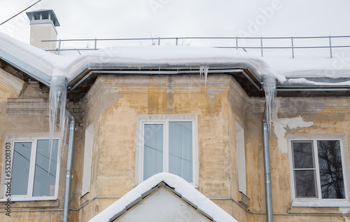 Long, frozen icicles hang on a drainpipe, spring or winter. Wall of an old brick house with windows. Large cascades of icicles in smooth, beautiful rows. Cloudy winter day, soft light.