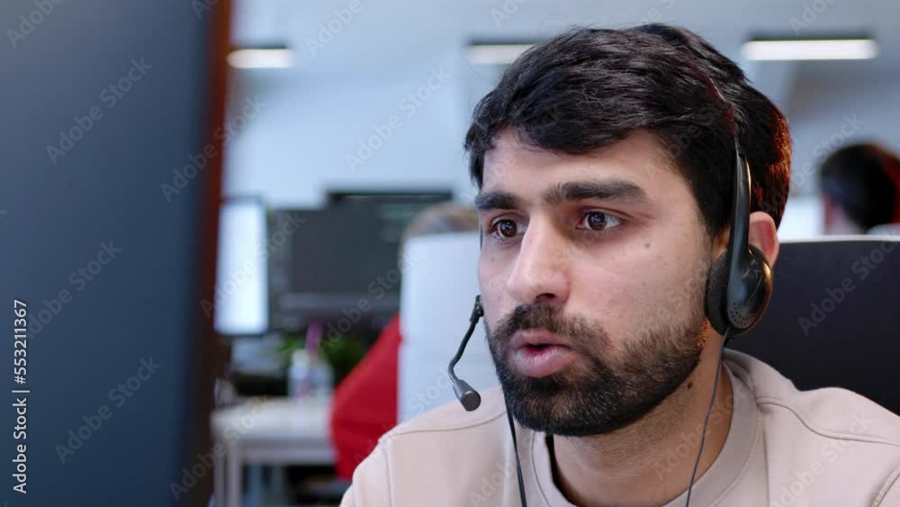 Close-up portrait of young handsome worker in call center talking with customers online wearing headphones sitting in office. Professional bearded man working in customer support sphere.
