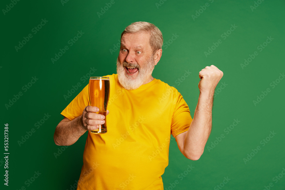 Portrait of senior man in yellow T-shirt emotionally posing with beer isolated on green background. Football fan Portrait of senior man in yellow T-shirt emotionally posing with beer isolated on green background. Football fan