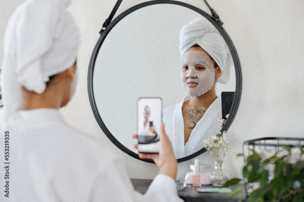 African American girl with hydrating sheet mask on face taking selfie in front of mirror while holding smartphone in front of herself African American girl with hydrating sheet mask on face taking selfie in front of mirror while holding smartphone in front of herself