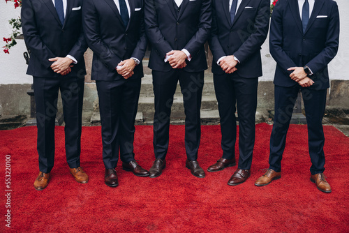 Wedding. Groom's friends. Business. A group of men in suits and leather shoes stand on a red carpet in front of the entrance to the building.