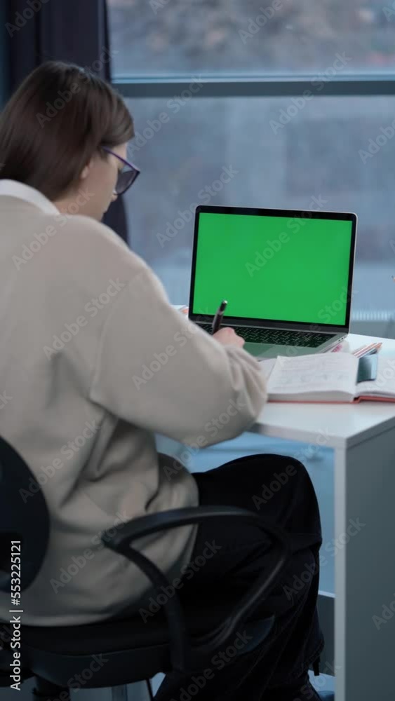 Woman Uses Laptop with Green Mock-up Screen. Girl Student Taking Notes on Notebook from Laptop at Online Lesson Woman Uses Laptop with Green Mock-up Screen. Girl Student Taking Notes on Notebook from Laptop at Online Lesson