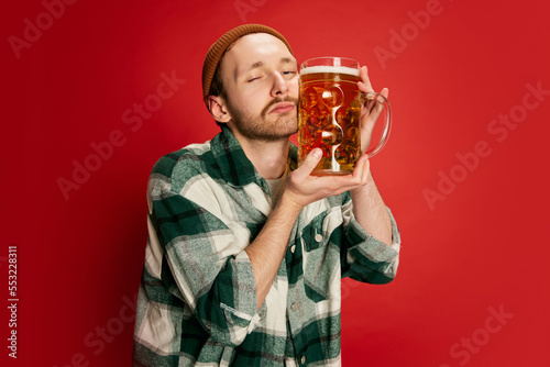 Foto Portrait of young man in casual checkered shirt posing with lager beer isolated over red background