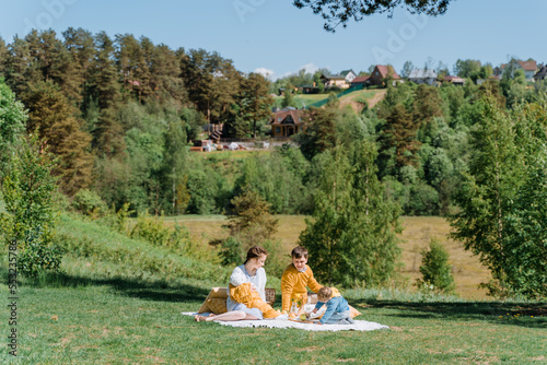 A young family with two children sits on a plaid on a country lawn under a large tree and enjoys a picnic in sunny weather, copyspace, yellow,blue and white colors © Елизавета Завьялова