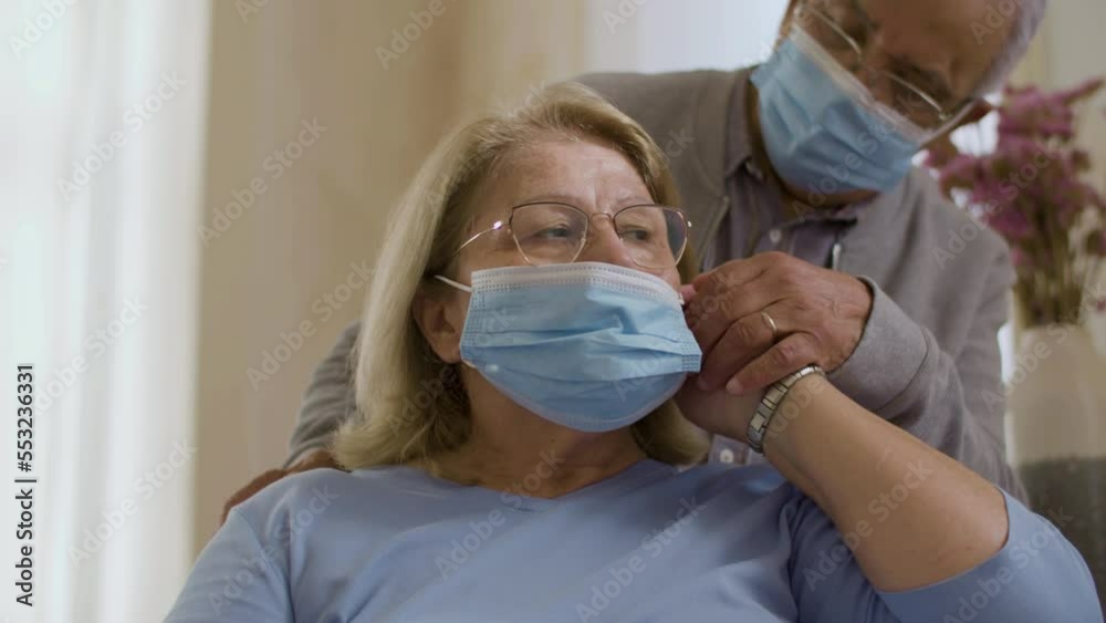 Medium shot of man helping wife putting on protective mask. Front view of senior couple sitting at home during self-isolation, taking care of health. Retirement, pandemic, coronavirus concept
