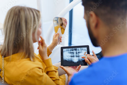 Rear view of dentist showing x-ray scan at digital tablet to his patient.
