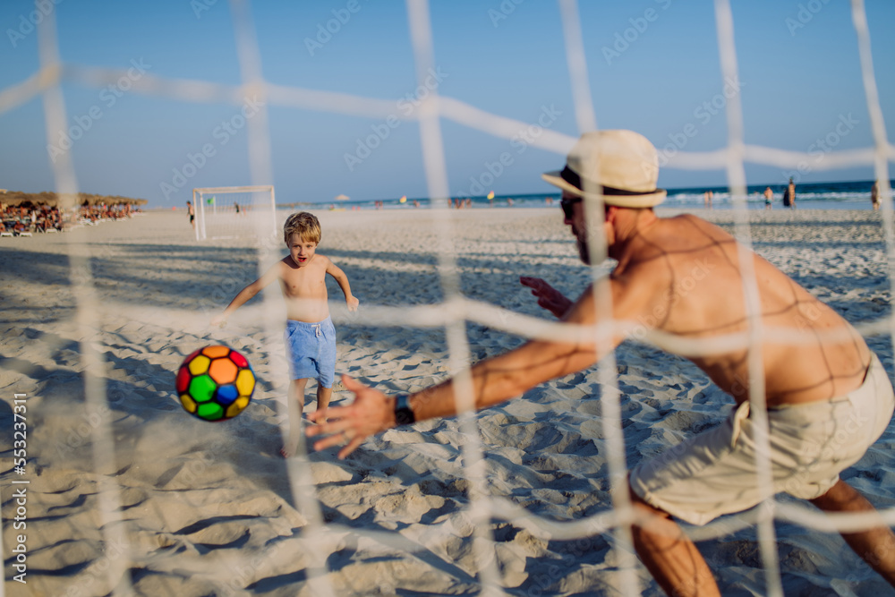 Father with his son plaing football on the beach. - obrazy, fototapety, plakaty 