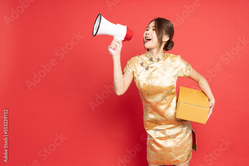Young Asian woman wearing golden traditional cheongsam qipao dress holding package parcel box and megaphone isolated on red background, Delivery courier and shipping service concept