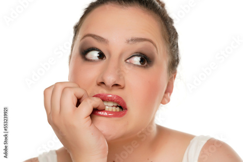 Young concerned chubby woman biting her fingernails on white background.
