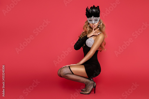 Elegant sexy woman in evening dress and carnival mask. Sensual glamour model in party dress and high heels on red background in studio. Vogue. Masquerade