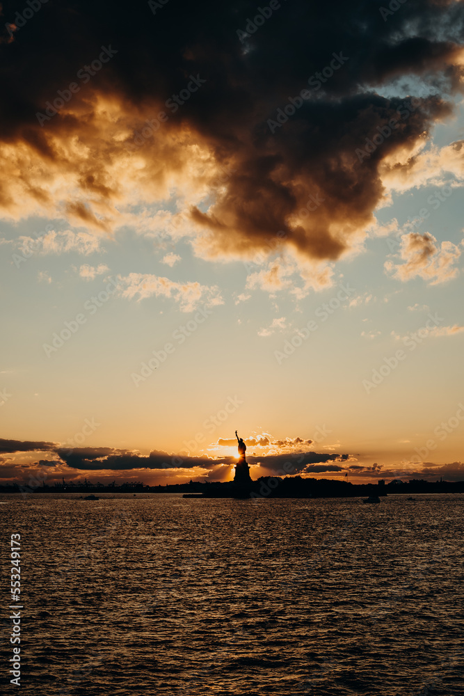 The picture shows the Statue of Liberty at sunset, with a warm glow over its copper surface and a colorful sky. The water surrounding the statue is calm.