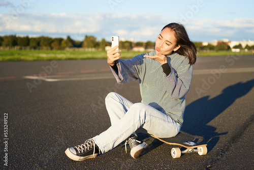 Beautiful korean girl takes selfie on smartphone, takes photo with her skateboard, enoys sunny day outdoors photo