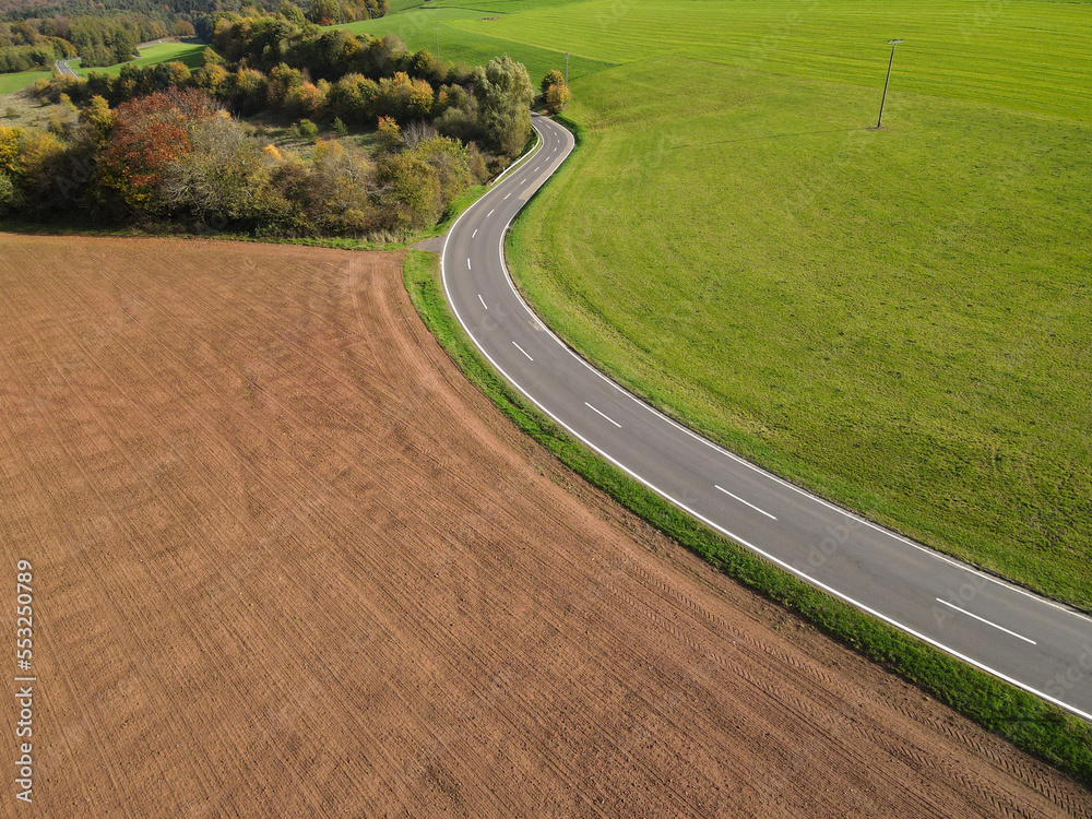 Aerial view of a long country road between plowed agricultural fields, trees and meadow on a warm day in autumn