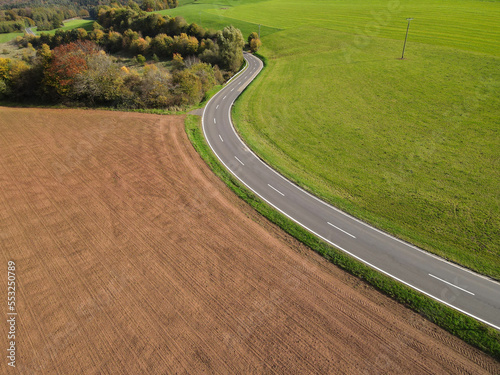 Aerial view of a long country road between plowed agricultural fields, trees and meadow on a warm day in autumn