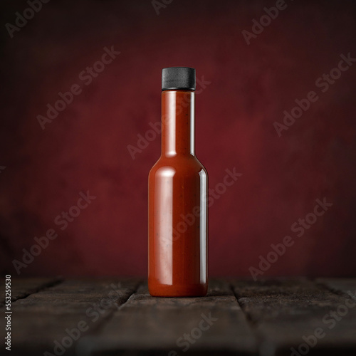 Glass bottle of red hot sauce on wooden surface. Chili pepper sauce. Ketchup. Tomato sauce. Vegetarian food. Photo for advertising. Dark red background. Copy space. Soft focus. Side view.