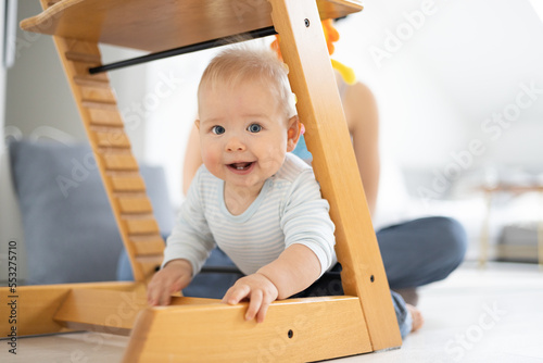 Happy infant climbing under traditional scandinavian designer wooden high chair and in modern bright home. Cute baby smiling in camera. photo