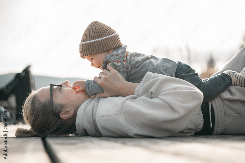 Happy family. Young mother playing with her baby boy infant oudoors on sunny autumn day. Portrait of mom and little son on wooden platform by lake. Positive human emotions, feelings, joy Happy family. Young mother playing with her baby boy infant oudoors on sunny autumn day. Portrait of mom and little son on wooden platform by lake. Positive human emotions, feelings, joy