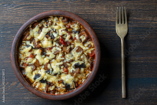 Stewed potato, carrot, onion, tomato, prunes and cheese in a clay bowl on wooden background, closeup, top view