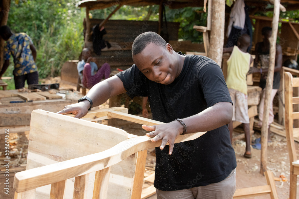 Young African carpenter searching and choosing wood and using sandpaper to rub the wooden plank at workshop table in carpenter factory