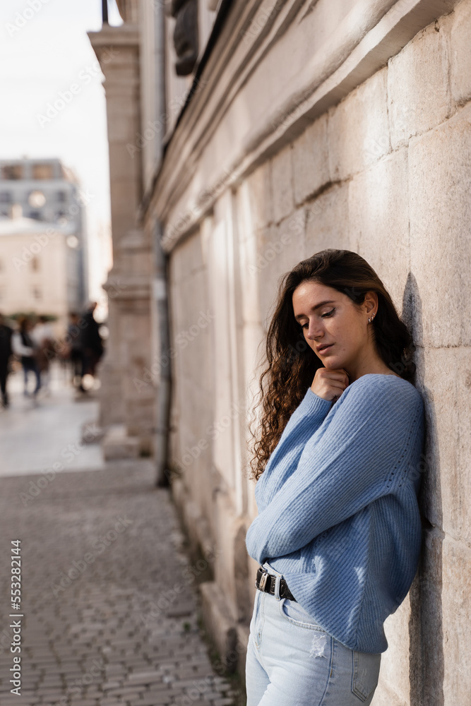 Portrait of attractive business woman with curly hair is posing on grey wall background. Confident girl model weared in blue pullover is posing outdoor in city. Portrait of attractive business woman with curly hair is posing on grey wall background. Confident girl model weared in blue pullover is posing outdoor in city.