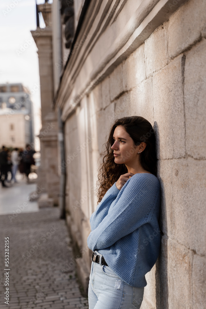 Confident girl model weared in blue pullover is posing outdoor in city. Portrait of attractive business woman with curly hair is posing on grey wall background.