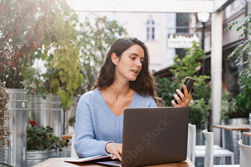 Busy business woman with laptop and phone is chatting and working remotely with colleagues. Cheerful girl with phone and laptop is working online with customers and chatting with team.