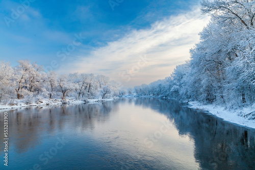 Beautiful view of the winter river. Trees against the background of the morning sky in a haze. Snow-covered trees after snowfall on the riverside are reflected in the water.