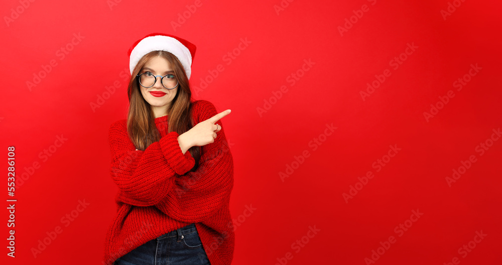 Fototapeta premium A beautiful young woman in a red sweater and santa hat points to an empty space for a discount. Christmas and New Year promotional offer.
