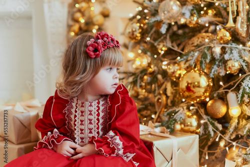 A little girl in a beautiful red dress is sitting near the Christmas tree and looking away