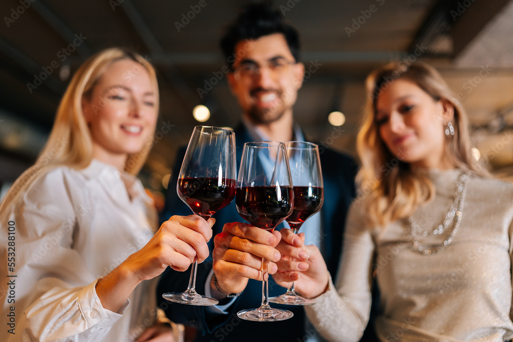 Focused on foreground of cheerful man in suit and eyeglasses and two pretty blonde women holding glasses of red wine, standing posing in restaurant. Happy young male and female enjoying nice dinner.