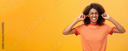 Waist-up shot of intense bothered dark-skinned woman clenchign teeth from discomfort and antipathy closing ears not hear botherind loud noise standing displeased and annoyed over orange wall photo