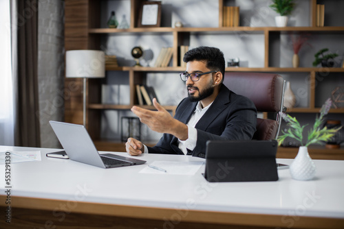 Confident businessman wearing suit and glasses sitting at desk with laptop during video chat, focused serious man working with new project, student studying online, research work
