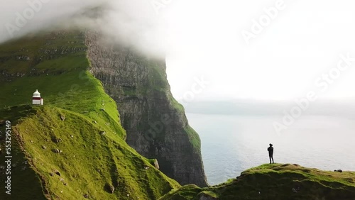 Aerial drone view of Kalsoy island with a man standing on a cliff, Faroe Islands. Amazing Faroese nature, green hills and beautiful rocky cliffs and mountain. Kallur lighthouse. Sunny day. 4k footage