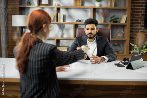 Back view of young red hair businesswoman giving bribe to smiling confident bearded businessman at workplace in modern office. Venality, bribe, corruption concept