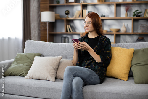 Attractive caucasian red hair woman sitting on couch and using modern smartphone indoors. Female freelancer taking break during working process at home.