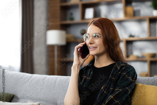 Close up portrait pf beautiful caucasian positive red hair woman sitting on couch and calling to her friend using smartphone. Smiling female freelancer taking break during working process at home.