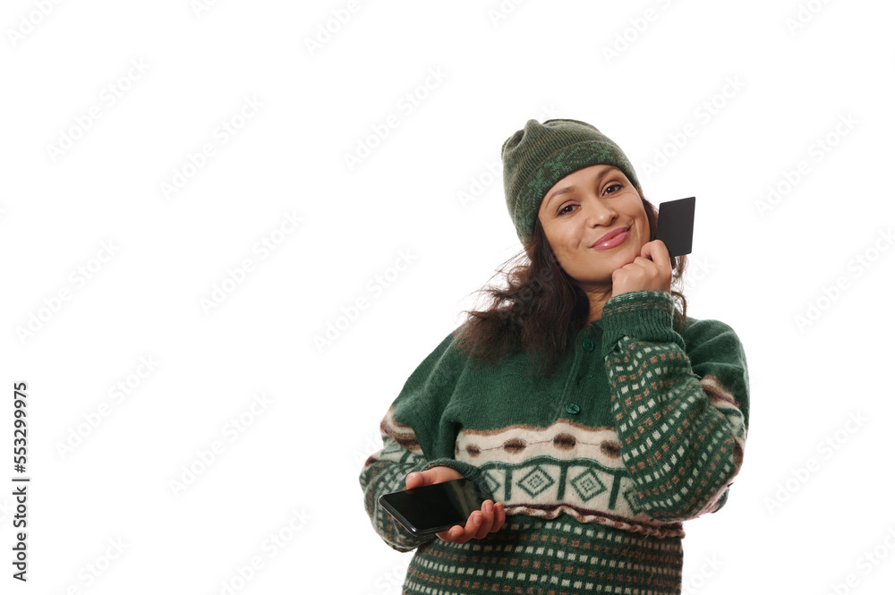 Isolated portrait on white background of charming woman in winter clothes, smiling looking at camera, holding smartphone and plastic credit card in her hands. Internet banking Online Shopping concept Isolated portrait on white background of charming woman in winter clothes, smiling looking at camera, holding smartphone and plastic credit card in her hands. Internet banking Online Shopping concept