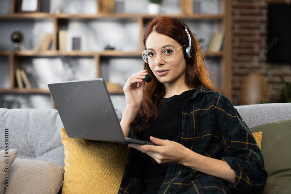 Video Conference. Smiling caucasian red hair woman having web call on laptop at home, talking at camera while sitting on sofa in living room.