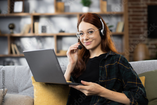 Video Conference. Smiling caucasian red hair woman having web call on laptop at home, talking at camera while sitting on sofa in living room.