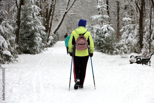Nordic walking at cold weather, healthy lifestyle. Woman with sticks walks in winter park on snow covered fir trees background