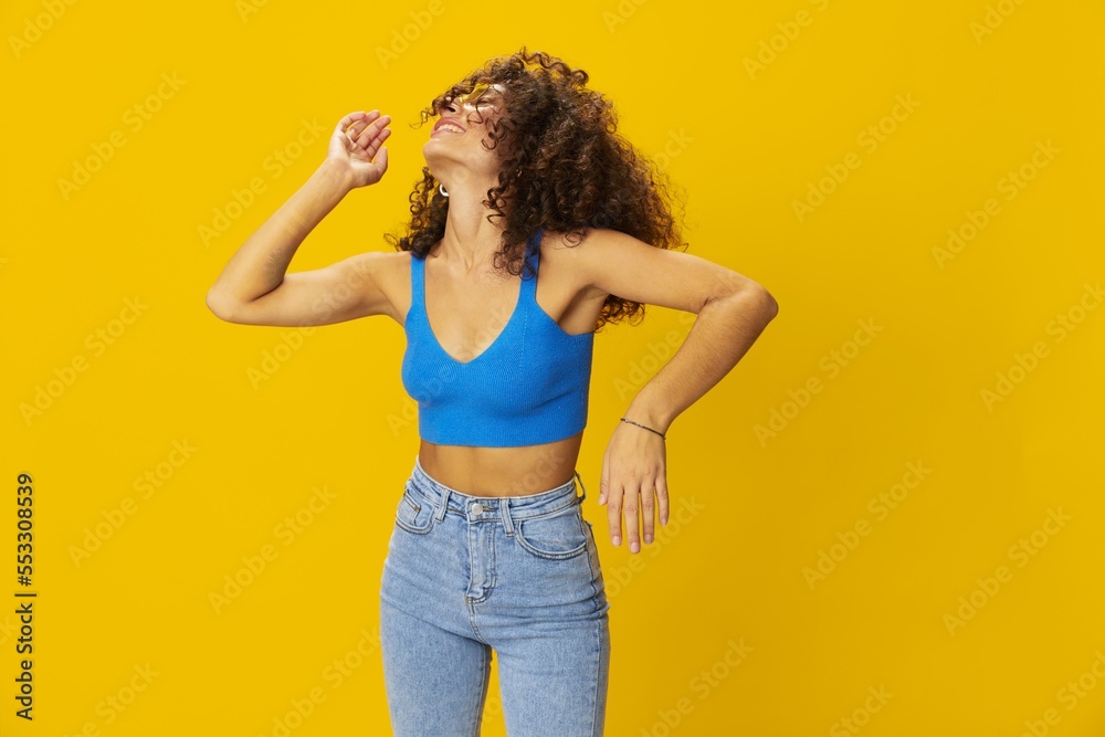 Woman with curly afro hair in a blue T-shirt on a yellow background dancing flying hair with sunglasses yellow, hand signs, look into the camera, smile with teeth and happiness, copy space Woman with curly afro hair in a blue T-shirt on a yellow background dancing flying hair with sunglasses yellow, hand signs, look into the camera, smile with teeth and happiness, copy space