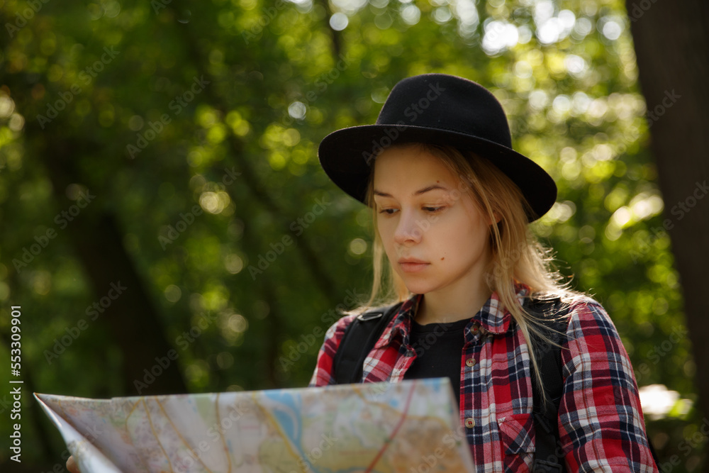 Obraz premium young caucasian woman in a hat looking at a geographic map in the shade of trees in a park or forest. High quality photo